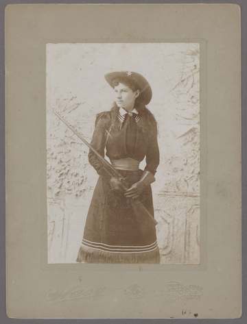 Three-quarter standing portrait of Annie Oakley cradling a rifle, wearing pearl earrings, wide-brimmed hat with star pin