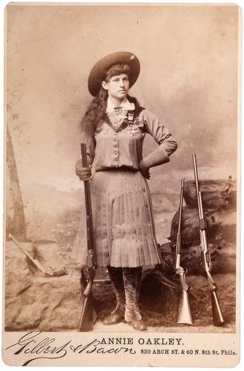Cabinet card showing Annie Oakley wearing shooting medals on her blouse, posed with a double-barrel Parker hammer shotgu