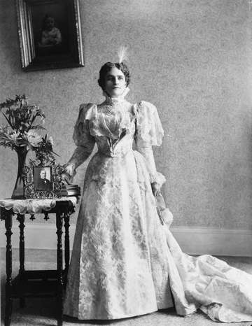 Ida S. McKinley, full-length portrait, standing, facing front. Picture of President McKinley on table beside her.
