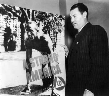 American boxer Jack Dempsey posed with Civil Defense poster "A Strong America" showing Jesse Owens on starting blocks.