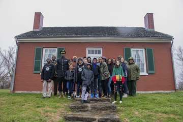Several children and adults stand in front of a red brick home with two chimneys and a staircase in the center foregroun